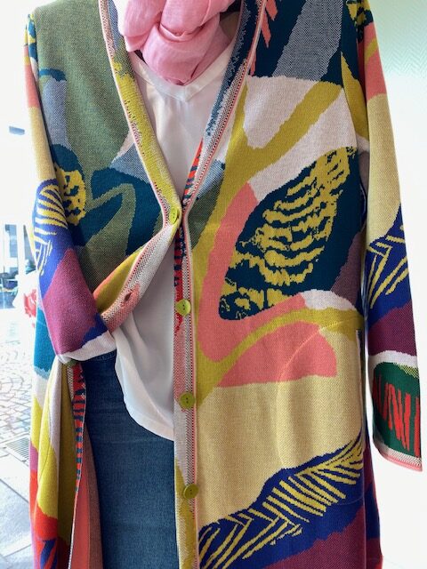Bunte Strickjacke mit abstraktem Muster, rosa Schal, weiße Bluse und blaue Jeans.