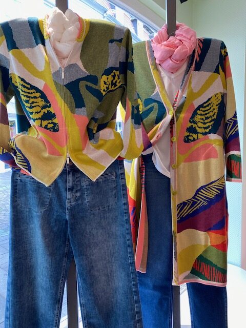 Zwei Schaufensterpuppen tragen bunte Cardigans und Jeans mit floralem Muster im Hintergrund.