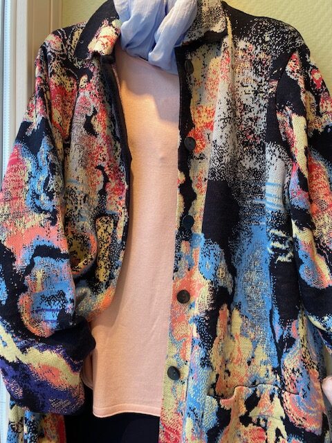 Bunte Jacke mit abstrakten Mustern, trägt jemand mit Schal und rosa Oberteil, angeschnitten.