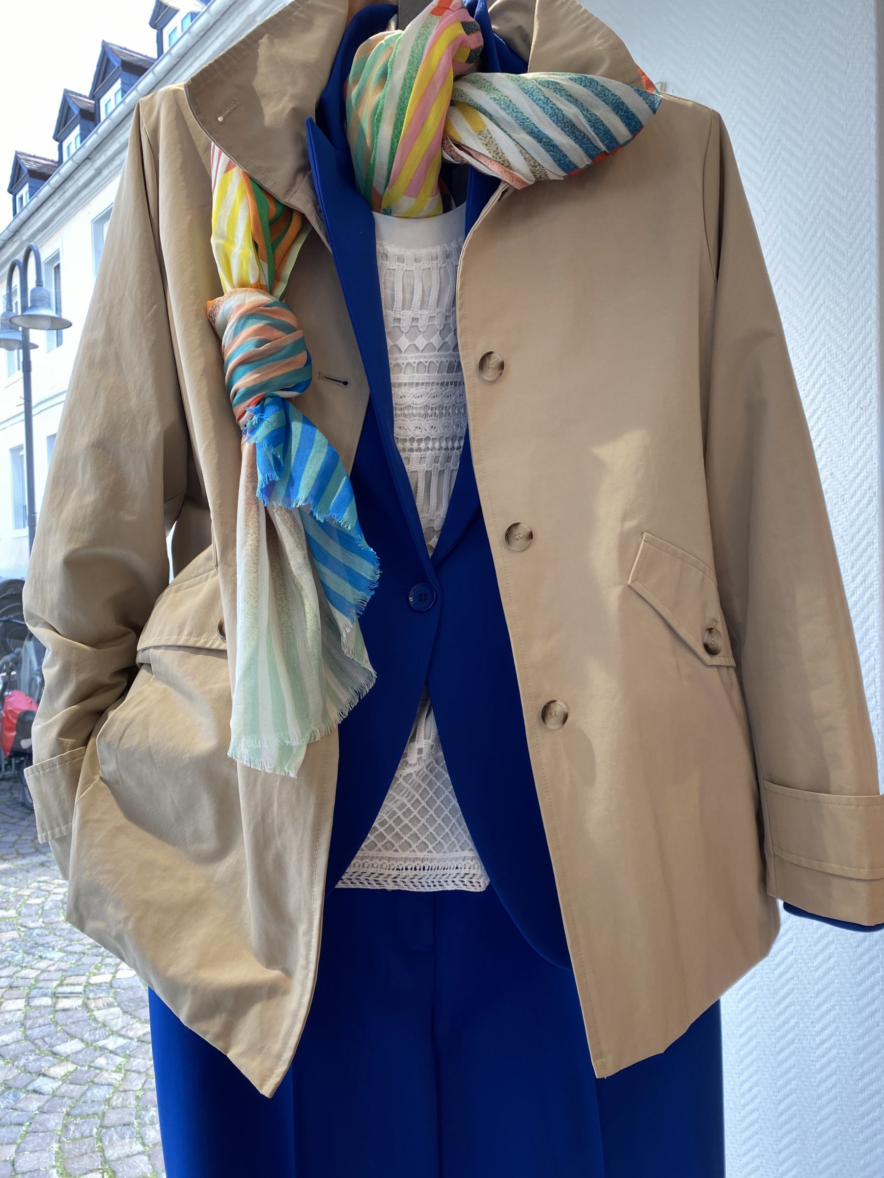 Beige Jacke mit bunter Schal über blauer Bluse, detailierter Spitzenstoff. Energetisch und stilvoll.
