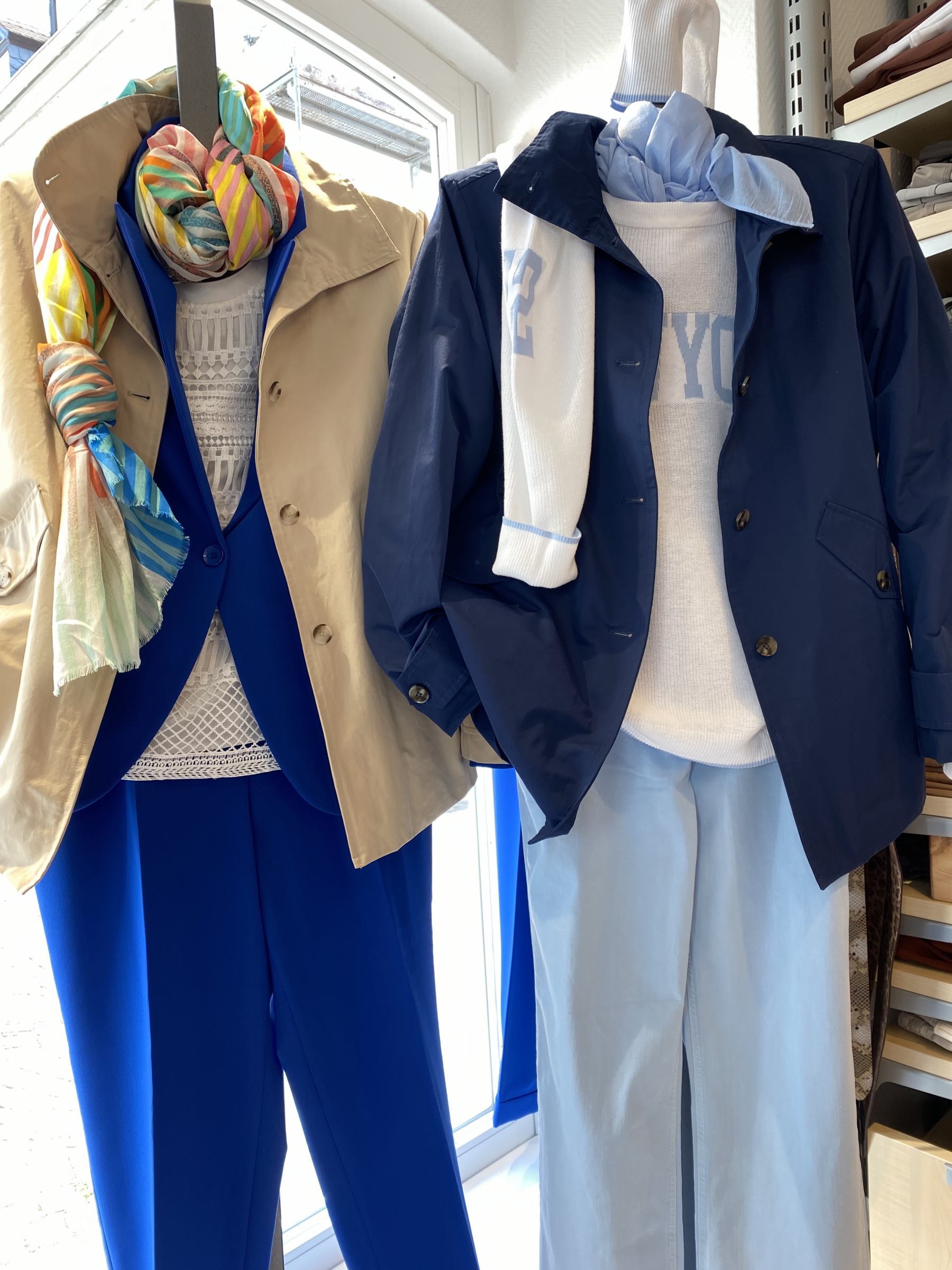 Zwei Schaufensterpuppen in bunten und blauen Outfits mit farbenfrohen Schals vor einem Fenster.