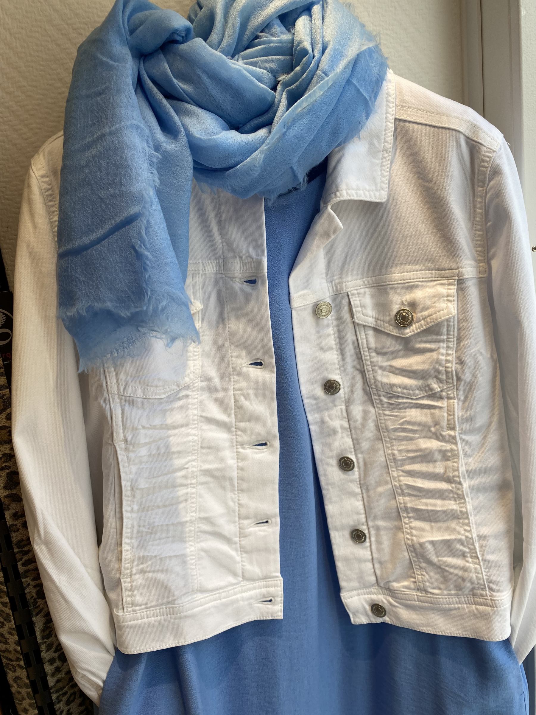 Weiße Jeansjacke mit blauer Schal und blauem Kleid auf Kleiderbügel.