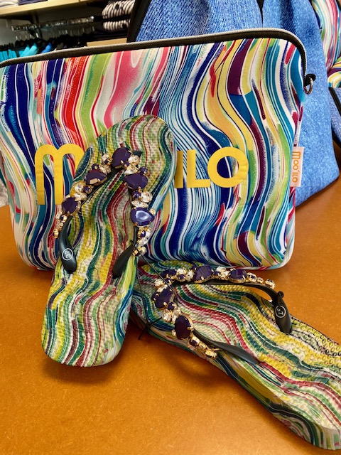 Bunte Flip-Flops mit Perlen vor einer farbigen Tasche mit Muster, auf einer braunen Fläche.