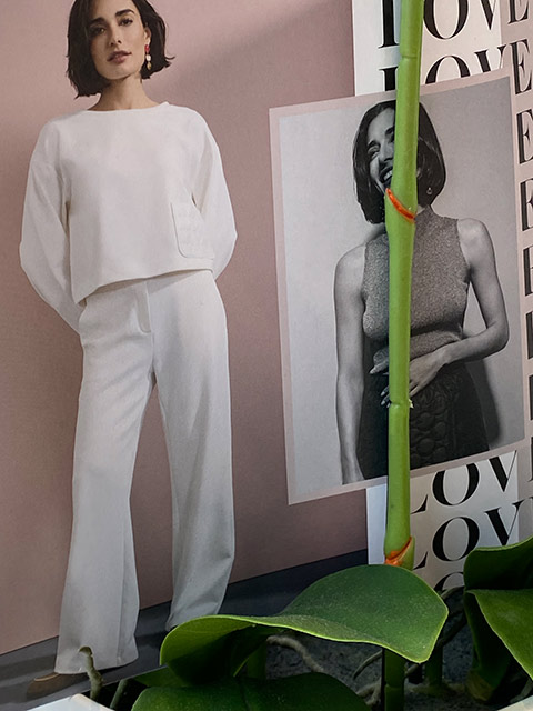 Frau in weißem Outfit, Ausdruck freundlich, hinter Grünpflanze und Bild.
