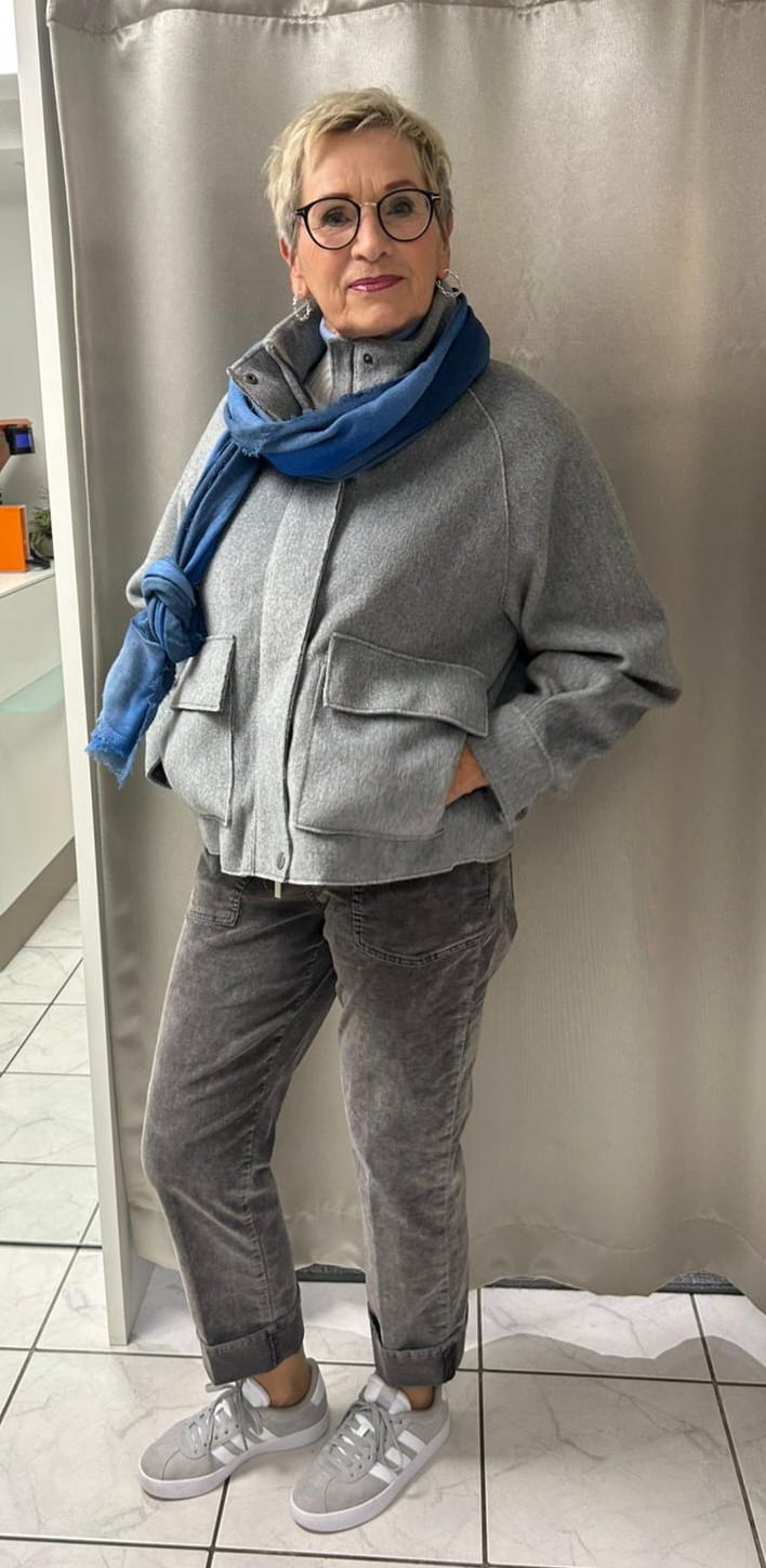 Eine ältere Dame mit kurzem Haar, Brille und grauer Kleidung lächelt freundlich