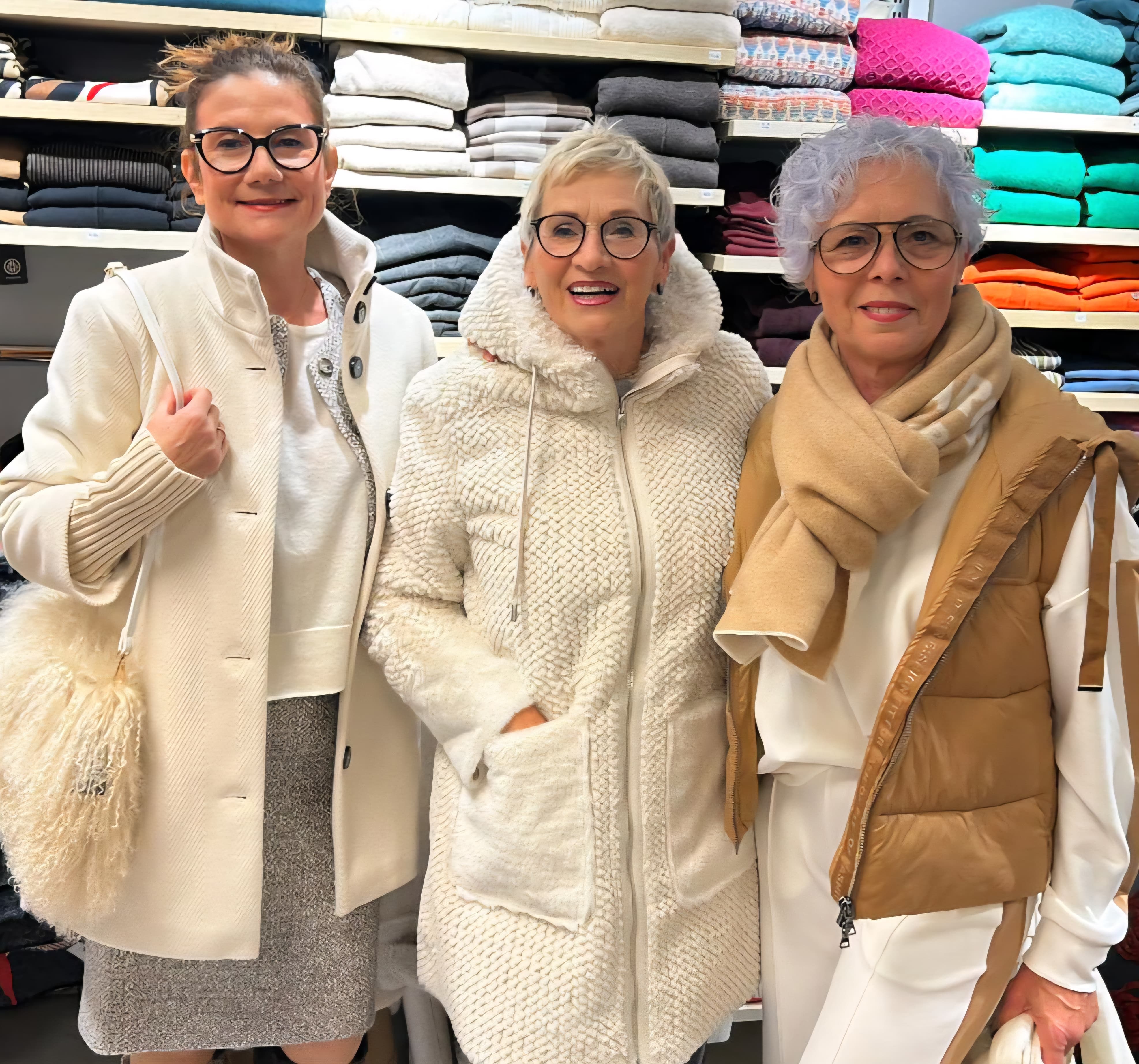 Drei stilvolle ältere Damen in heller eleganter Kleidung lächeln fröhlich vor farbigen Pullovern