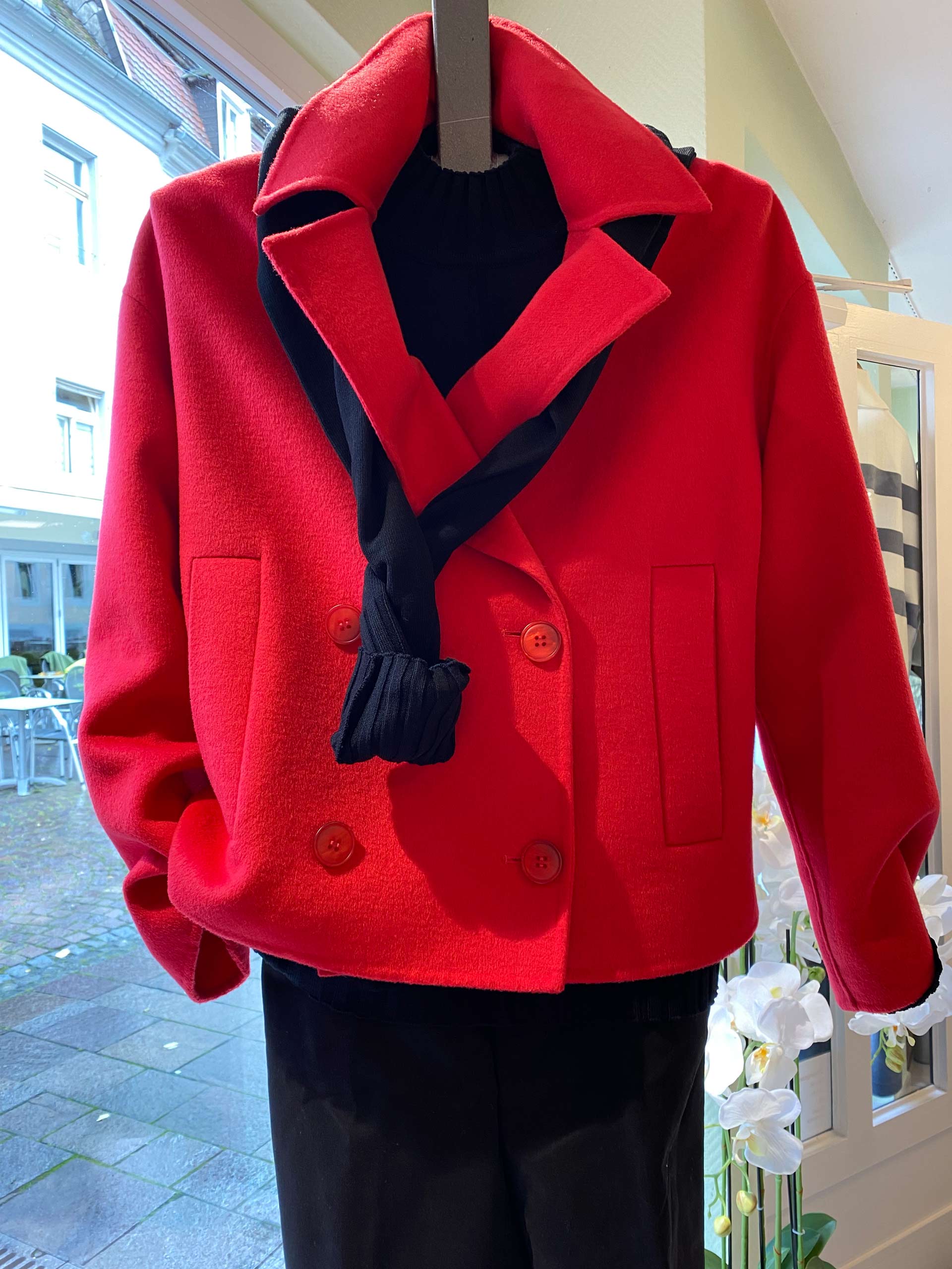 Ein roter Mantel mit doppelter Knopfreihe und schwarzem Schal auf einem Bügel