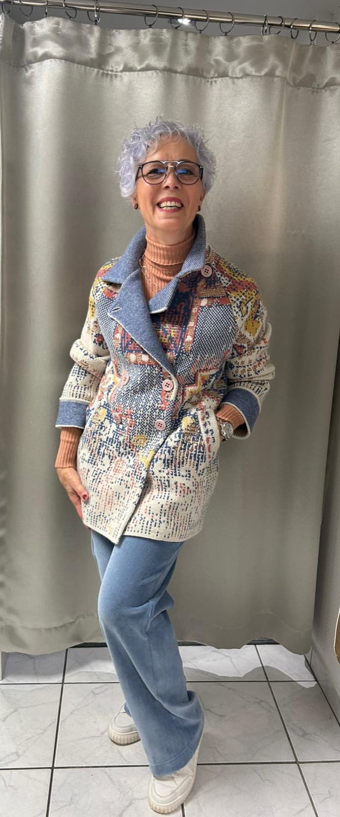 Eine lächelnde ältere Frau mit grauen Locken trägt bunte Strickjacke und blaue Hose