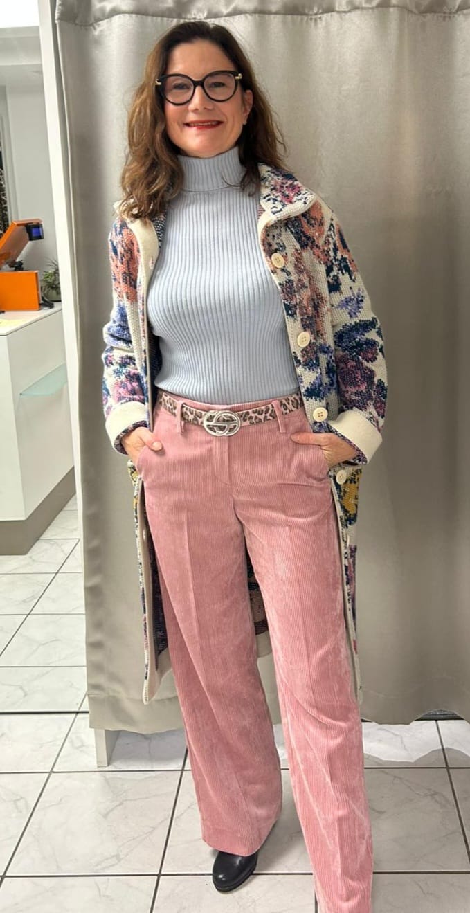Frau mit Brille lächelt freundlich und trägt bunte Jacke mit Cordhose in Rosa
