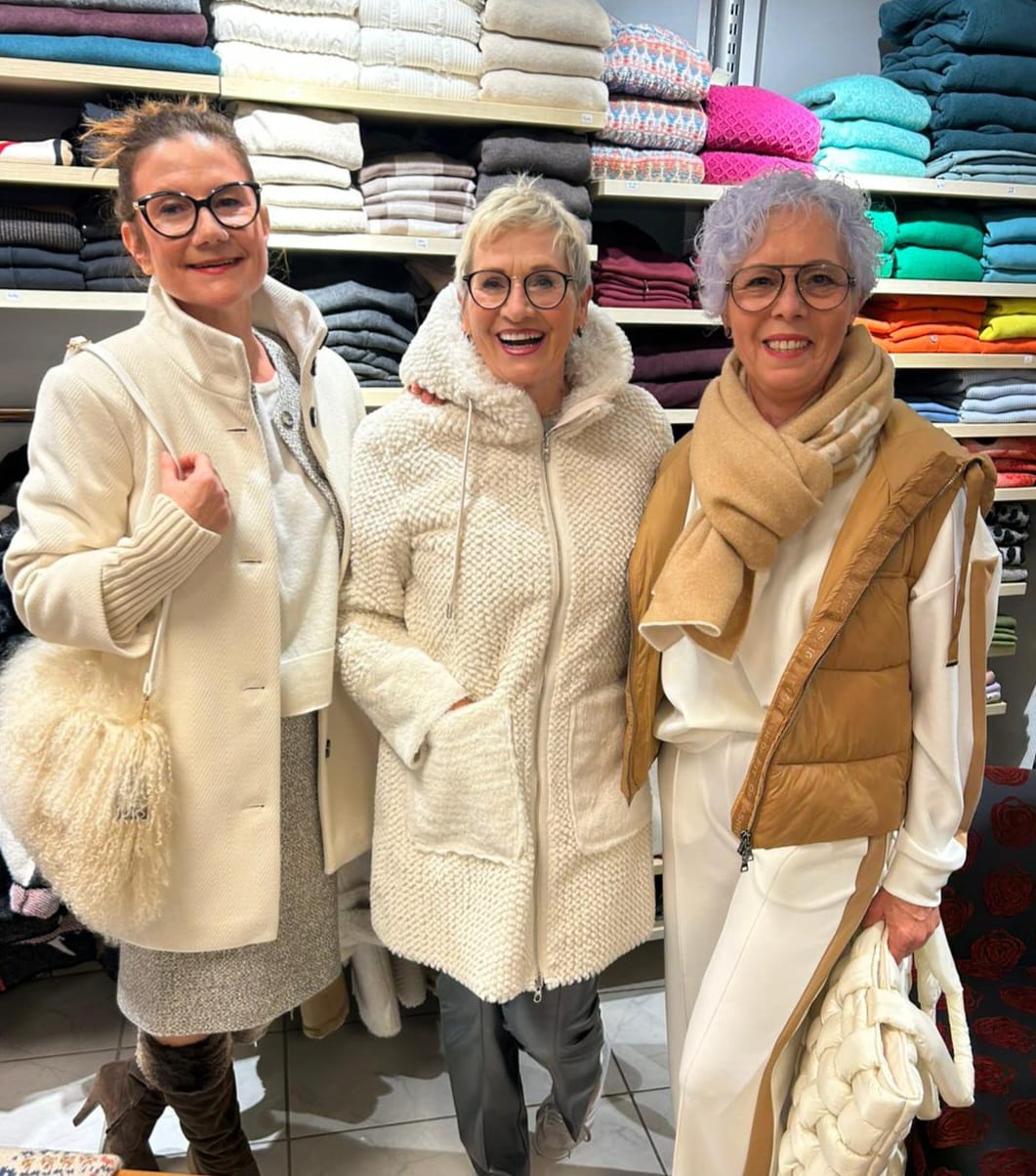 Drei ältere Frauen lächeln freundlich vor gefalteten bunten Handtüchern in einem Geschäft