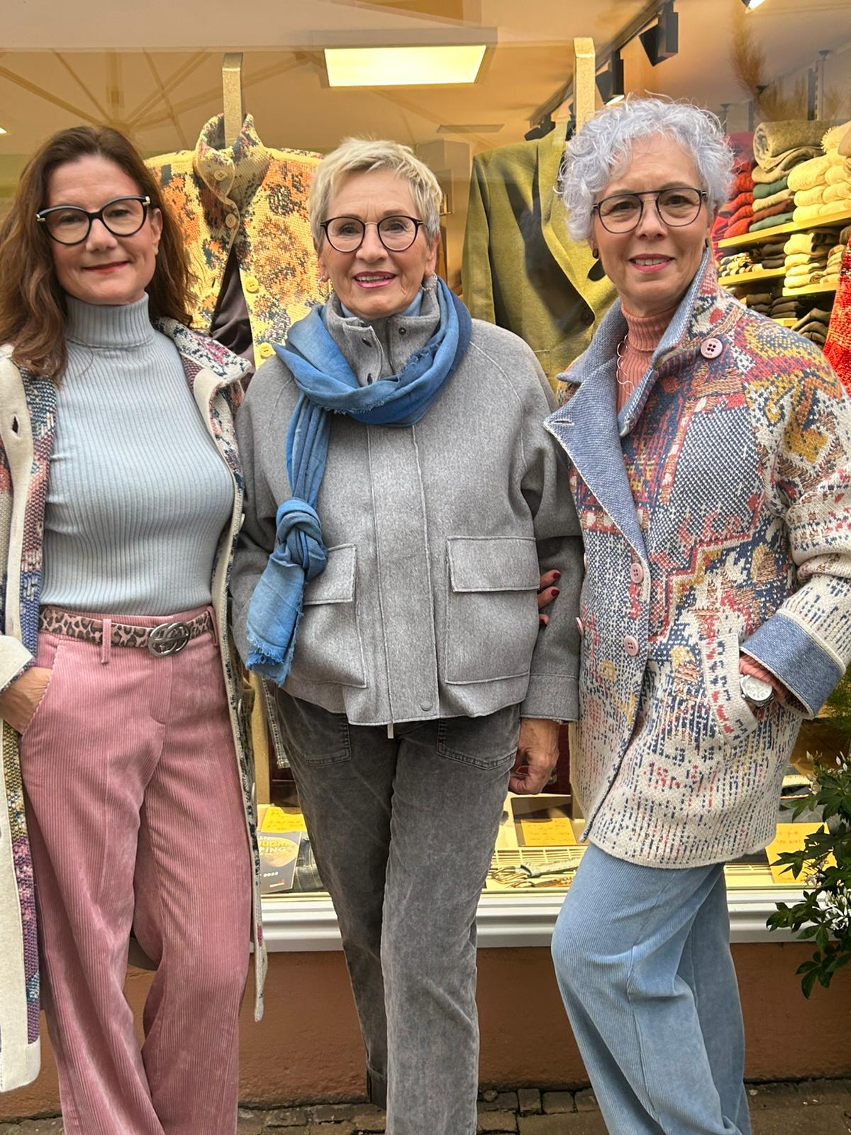 Drei ältere Frauen mit Brillen lächeln freundlich vor einem bunten Ladenfenster