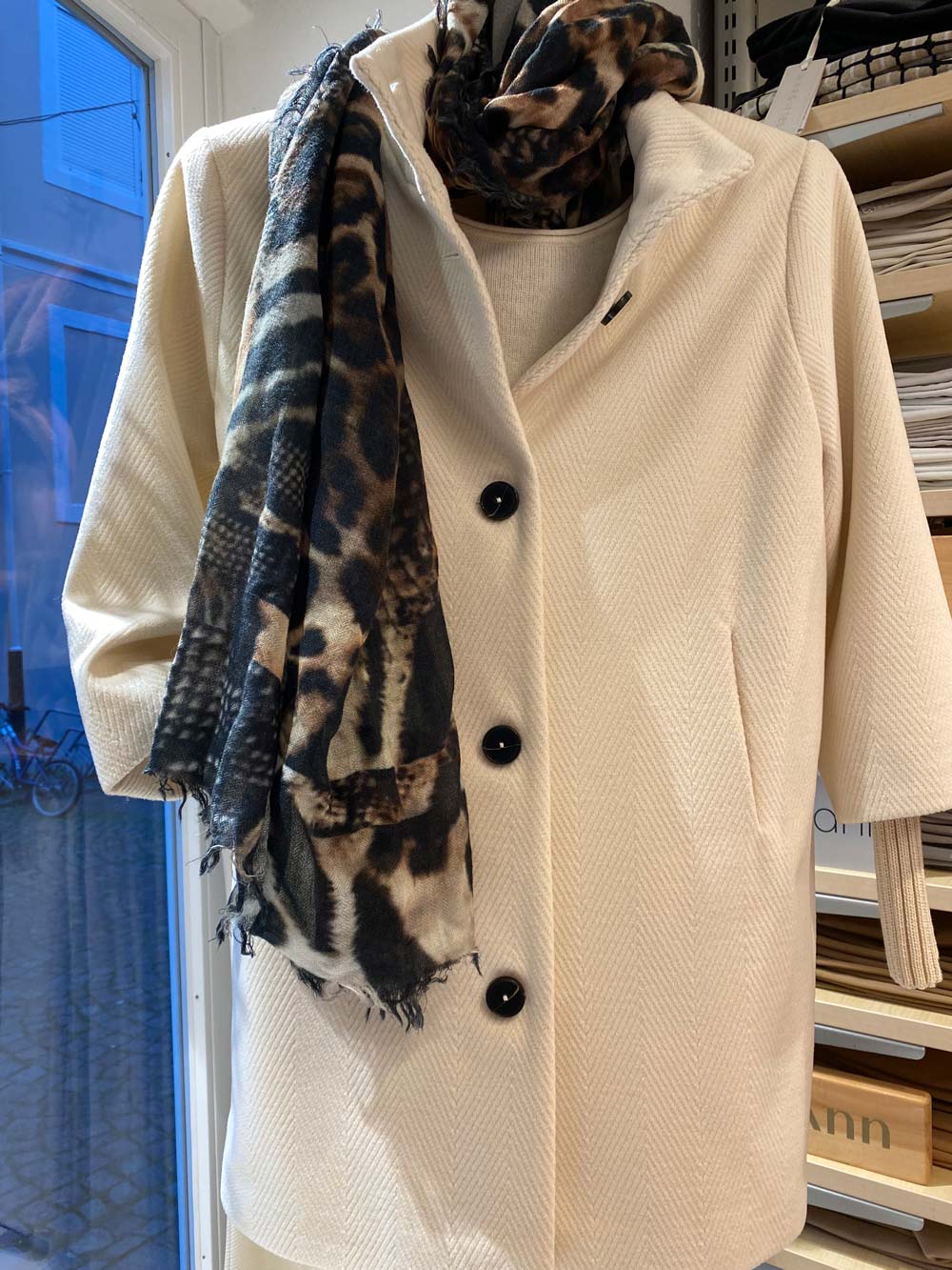 Beiger Mantel mit schwarzen Knöpfen und gemustertem Schal auf Kleiderbügel