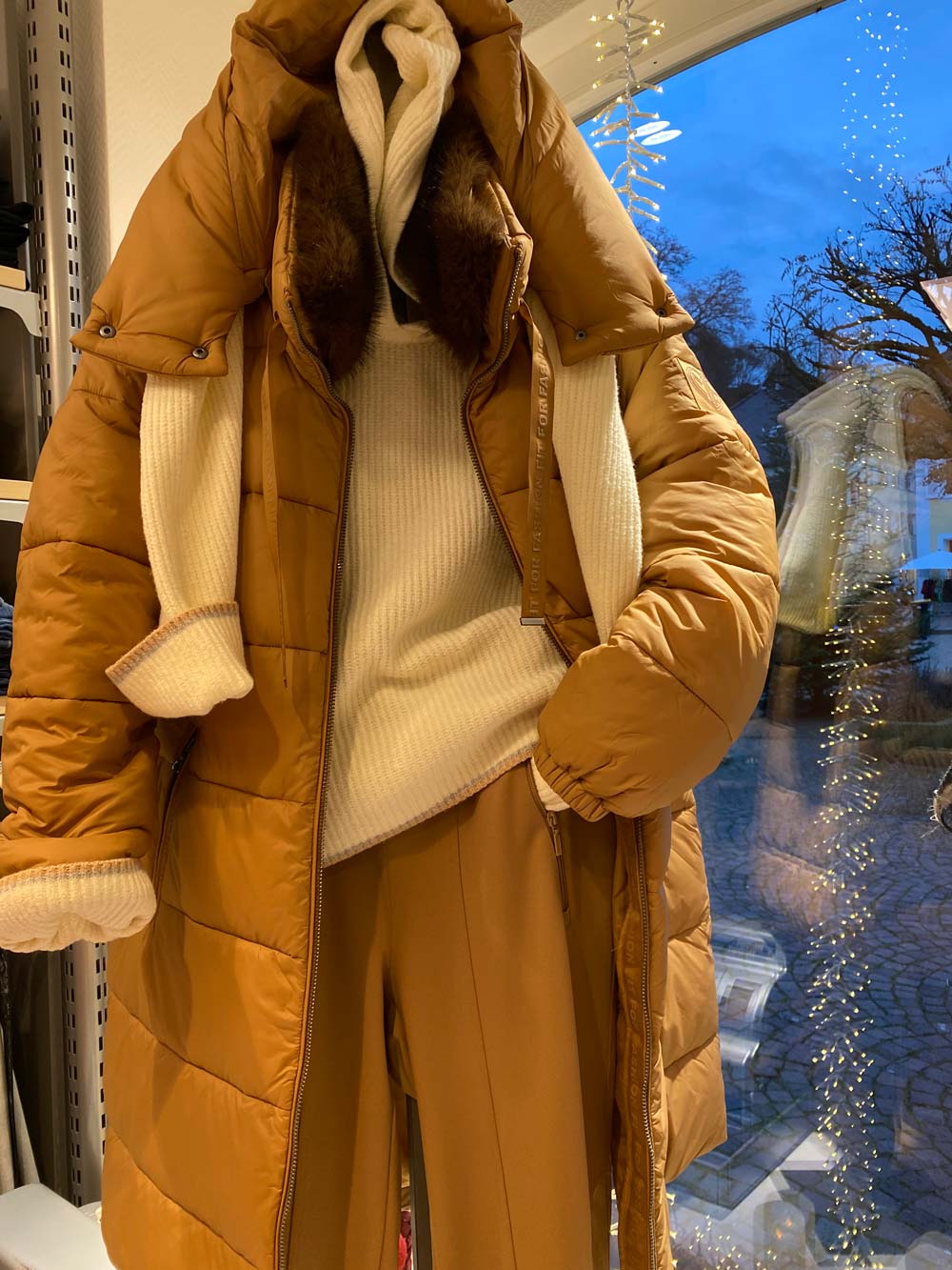 Eine Schaufensterpuppe trägt einen braunen Mantel, cremefarbenen Pullover und Schal