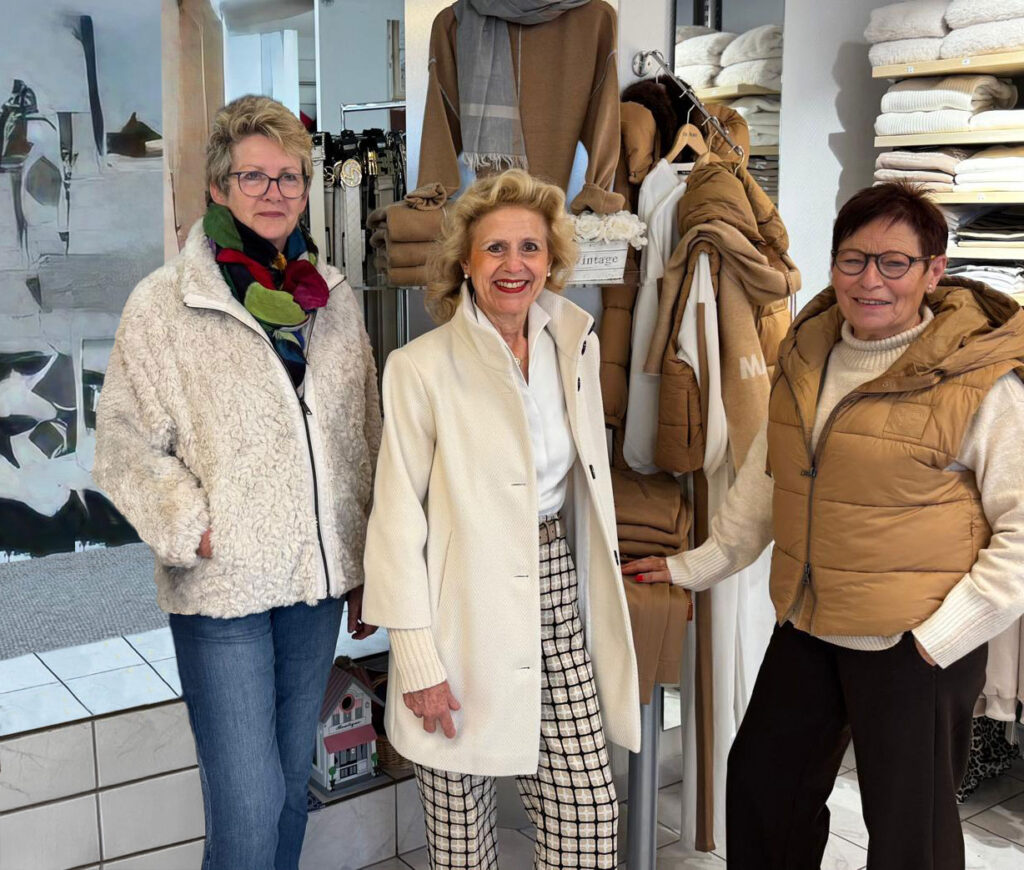 Drei ältere Damen mit freundlichen Gesichtern stehen vor beigen Kleidung und Handtüchern