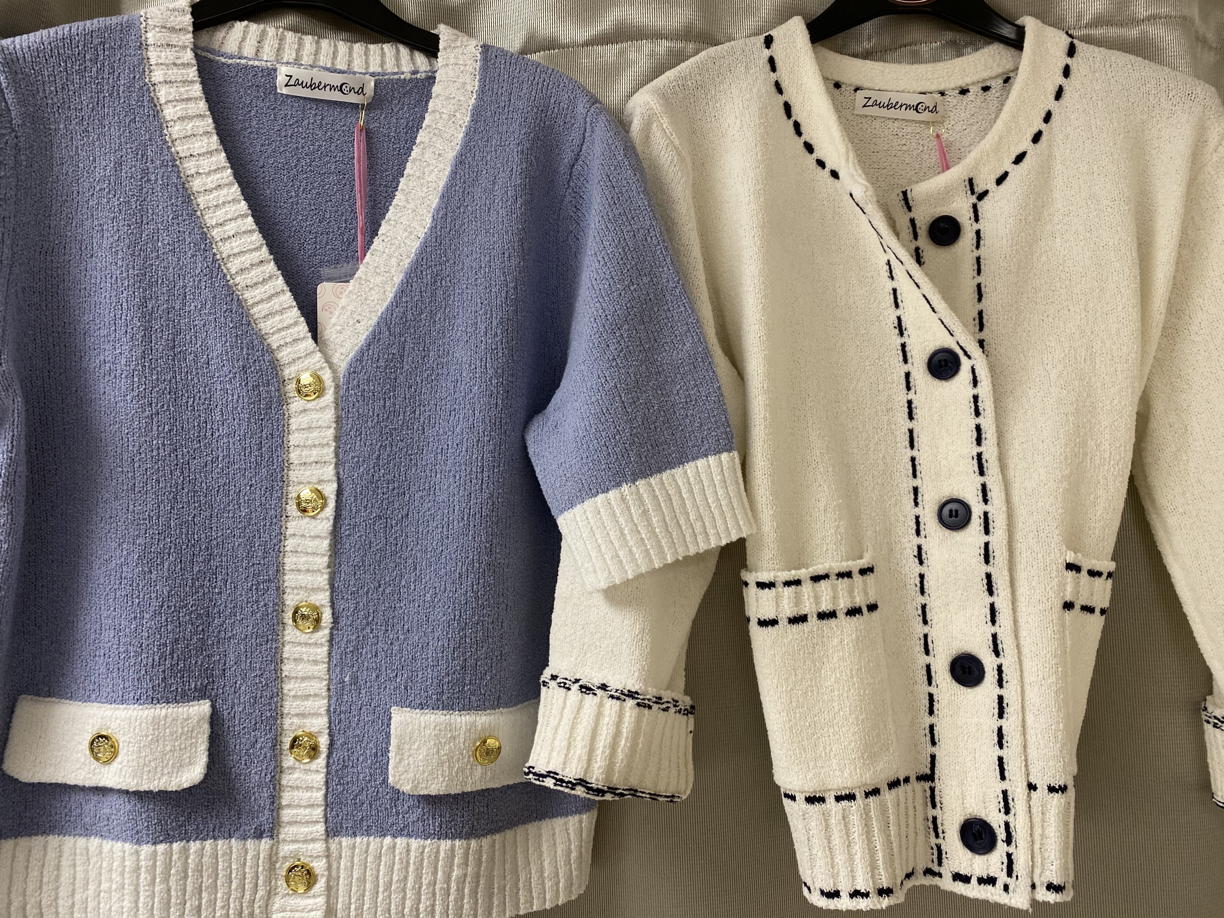 Zwei gestrickte Damenjacken, eine in Hellblau mit weißen Details und eine in Creme mit blauen Ziernähten, hängen nebeneinander und zeigen goldene Knöpfe sowie strukturierte Oberfläche
