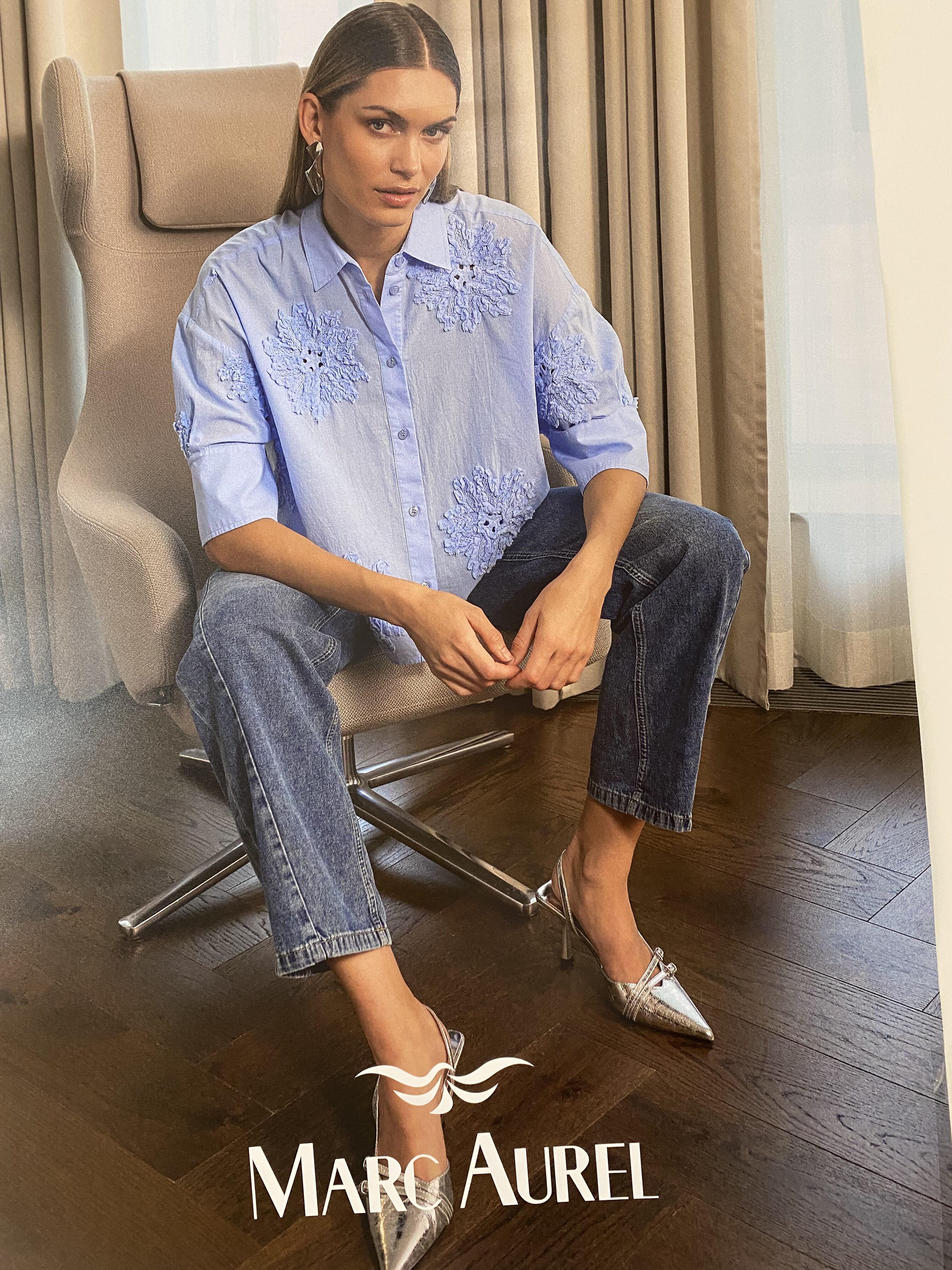 Eine Frau mit neutralem Gesichtsausdruck trägt eine hellblaue Bluse mit Blumenmuster und Jeans sowie silberfarbene Schuhe und sitzt auf einem modernen Stuhl vor einem Vorhang