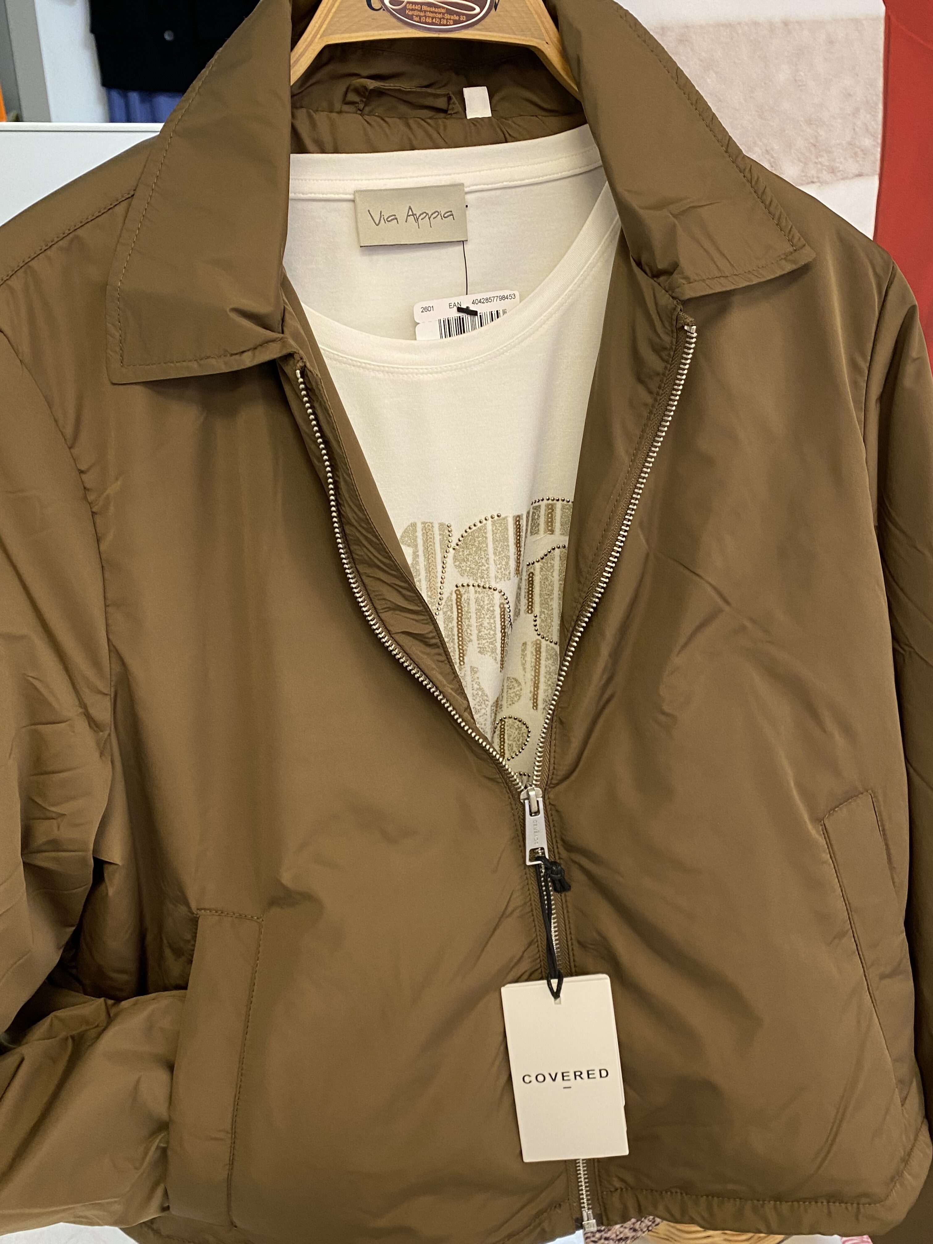 Eine braune Jacke mit Reißverschluss hängt auf einem Bügel über einem beigen Shirt