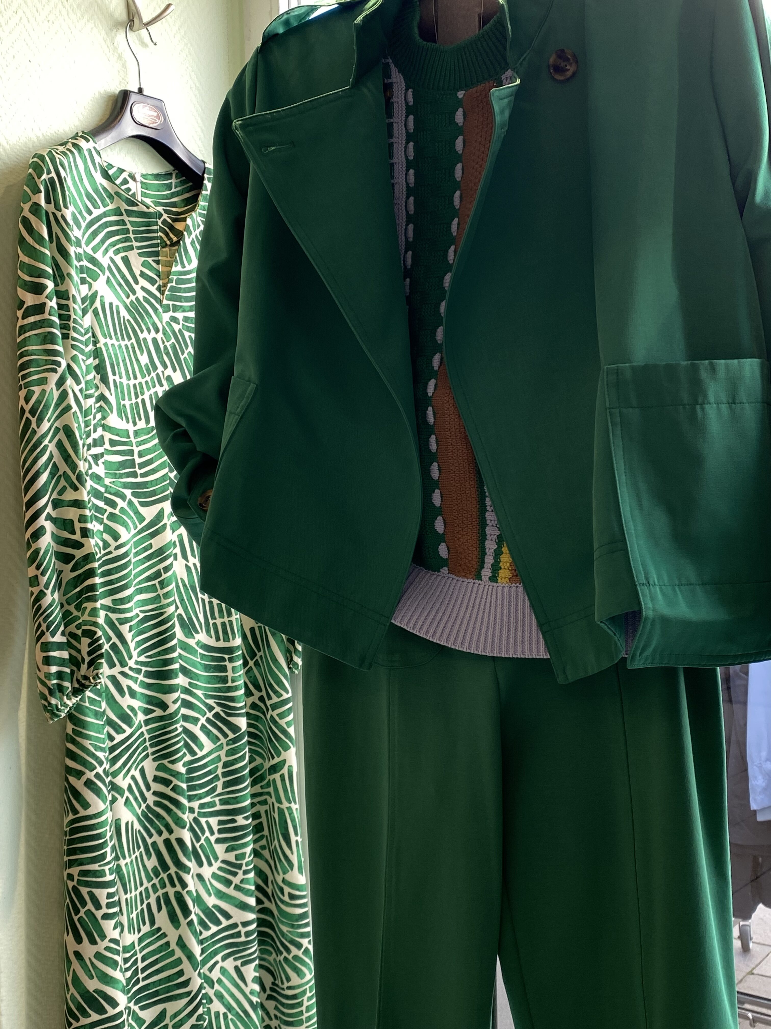 Ein grünes Outfit mit Mantel und Hose neben einem gemusterten Kleid in ähnlichen Farbtönen