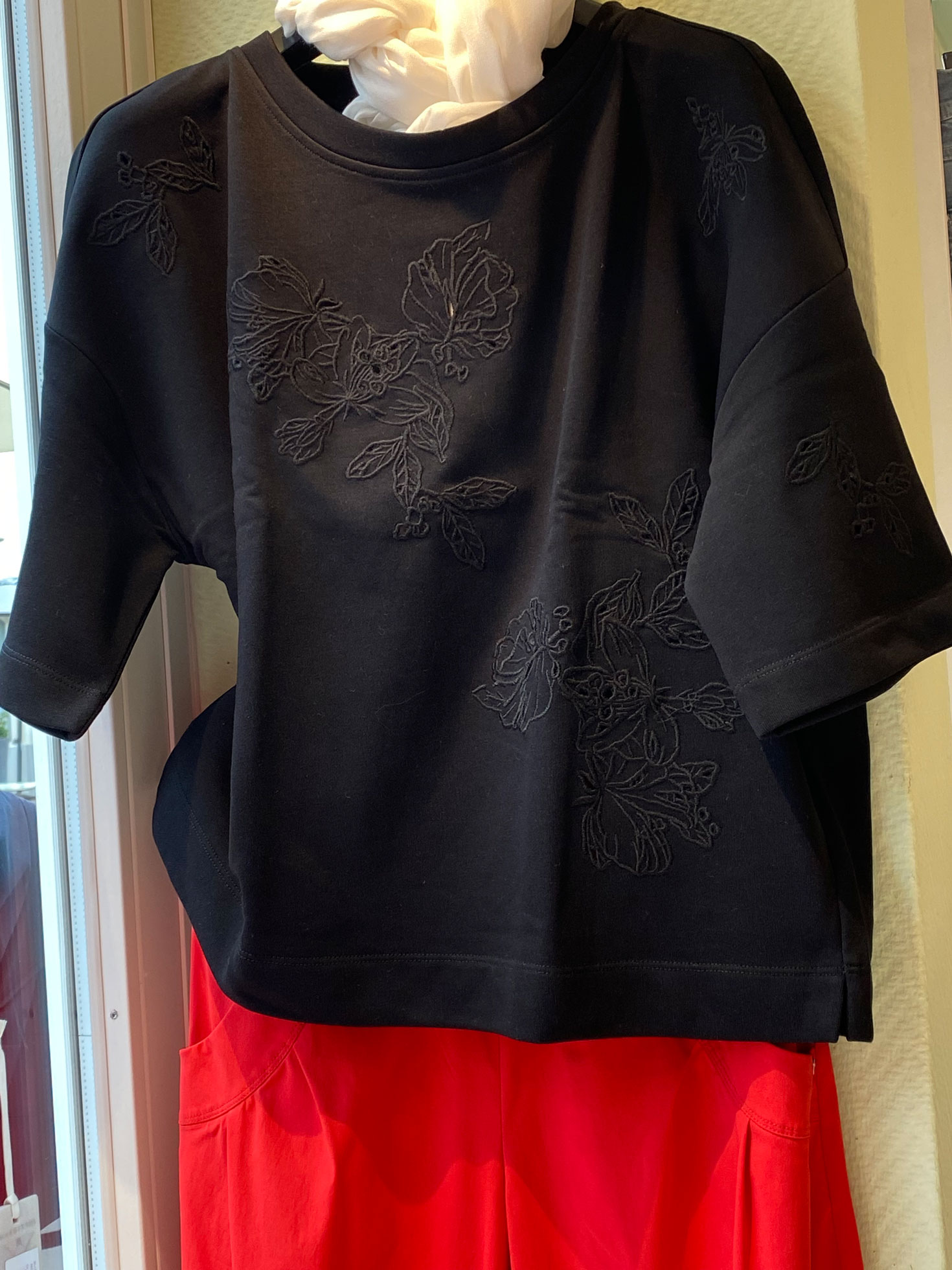 Schwarzes Sweatshirt ohne Person, schlicht, mit floralem Stickmuster, am Kleiderbügel über roter Hose