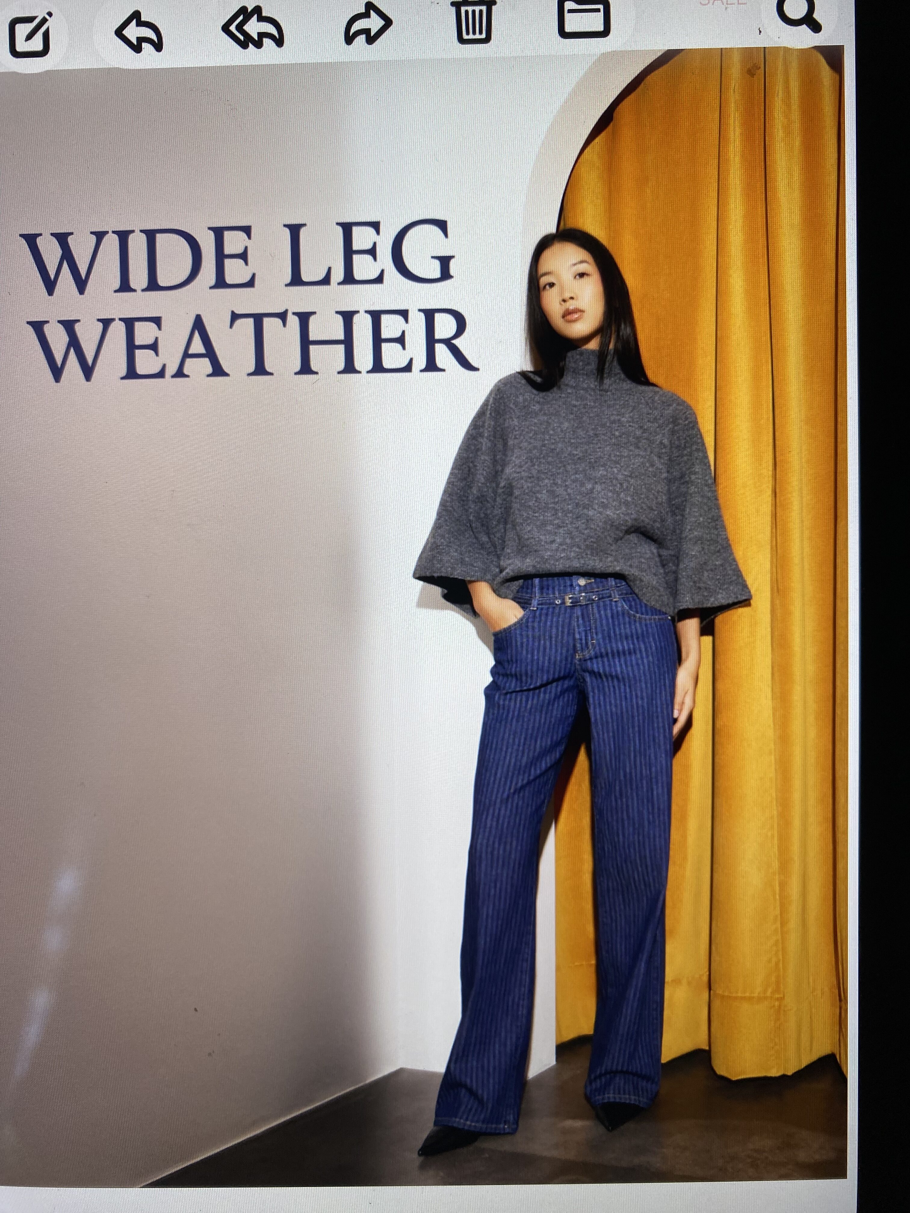 Junge Frau in grauem Pullover und weiter Jeans vor gelbem Vorhang