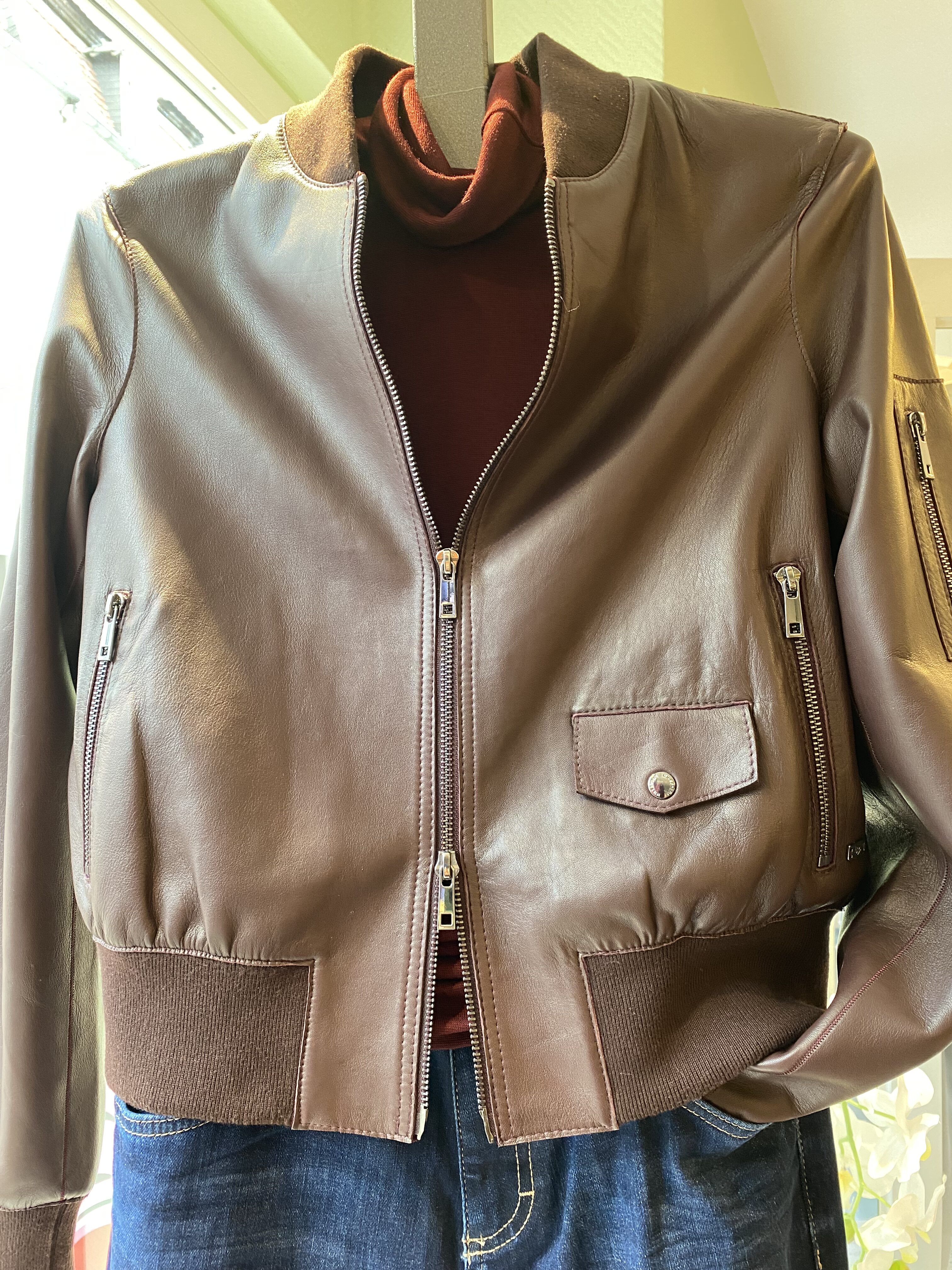 Braune Lederjacke mit Reissverschluessen ueber rotem Pullover und blauer Jeans